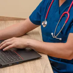 Lekarz w niebieskim fartuchu z czerwonym stetoskopem pisze na laptopie, szukając informacji, jaki lekarz zarabia najwięcej.