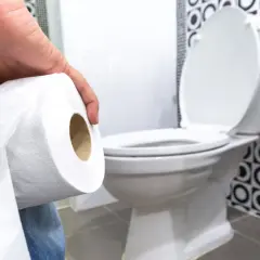 Dłoń trzymająca rolkę papieru toaletowego, w tle biała muszla klozetowa. Ktoś zmaga się z problemami z wypróżnianiem.